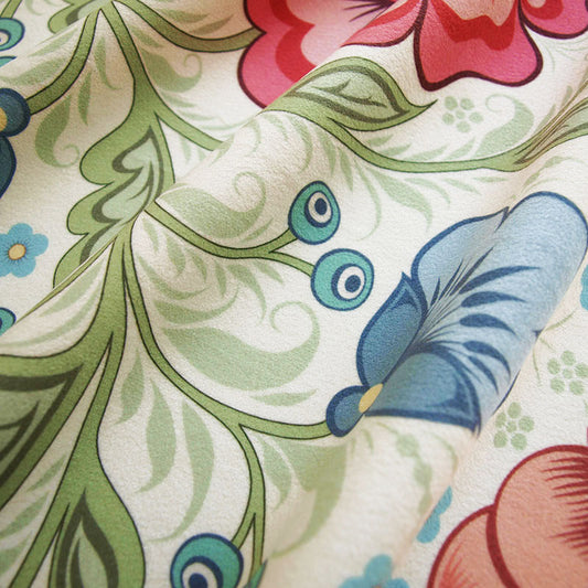 A close-up of a draped fabric with a floral and pattern featuring green leaves, blue and pink flowers, and teal circular accents on an off-white background.